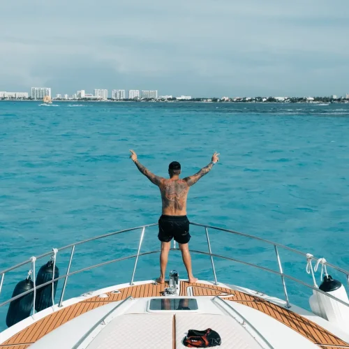 Tattooed man stands with arms raised on yacht bow overlooking Cancún.